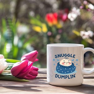 May include: White ceramic mug with the text "SNUGGLE DUMPLIN'" in blue, featuring a cartoon baby in a blue swaddle. The mug is on a wooden surface with pink and white tulips in the foreground and a blurred background of greenery and flowers.