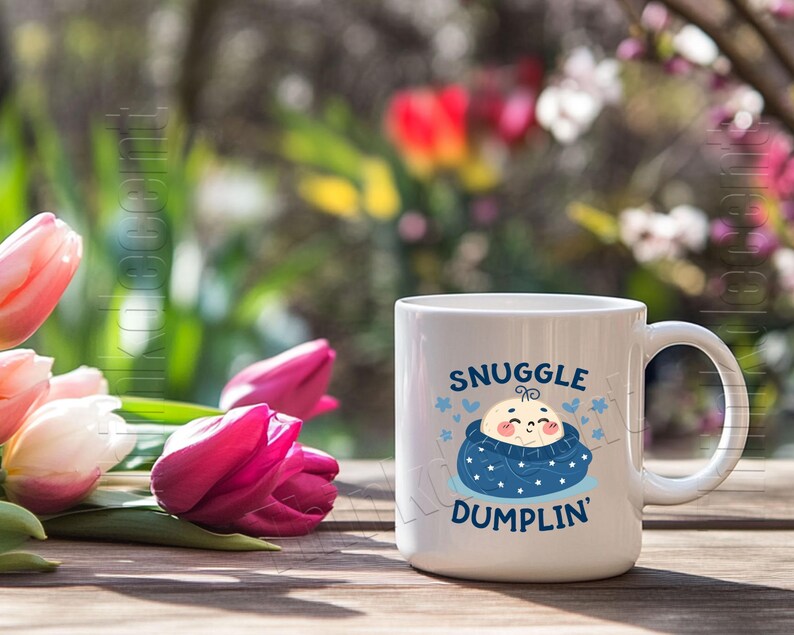 May include: White ceramic mug with the text "SNUGGLE DUMPLIN'" in blue, featuring a cartoon baby in a blue swaddle. The mug is on a wooden surface with pink and white tulips in the foreground and a blurred background of greenery and flowers.