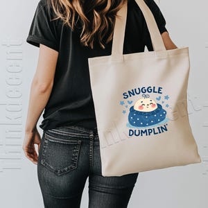 May include: Cream-colored tote bag with the words "Snuggle Dumplin'" and a cartoon baby dumpling design in blue and peach. The bag has two handles and is being held by a person wearing a black shirt and jeans.