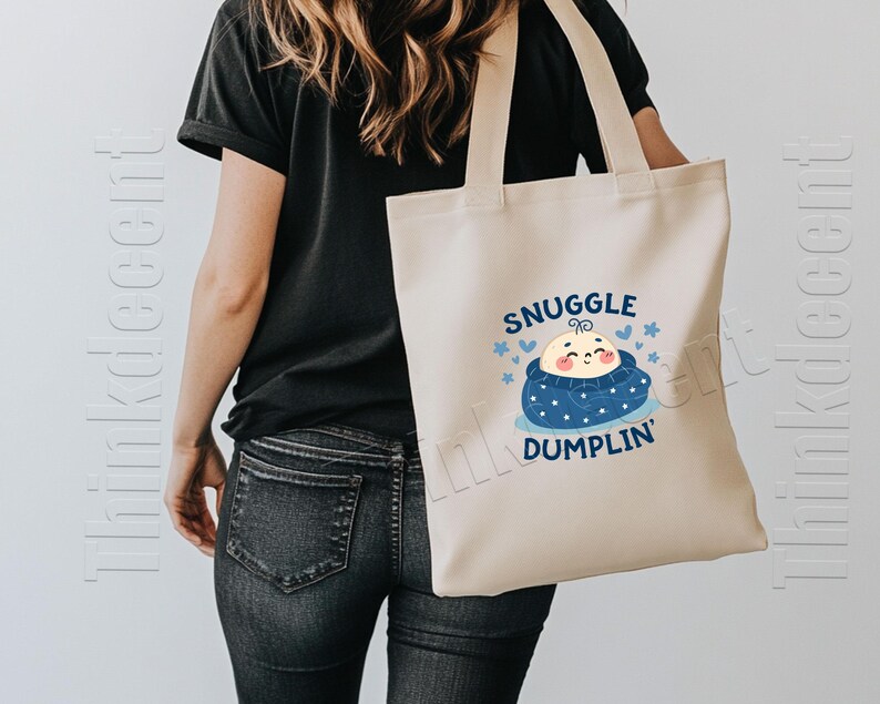 May include: Cream-colored tote bag with the words "Snuggle Dumplin'" and a cartoon baby dumpling design in blue and peach. The bag has two handles and is being held by a person wearing a black shirt and jeans.