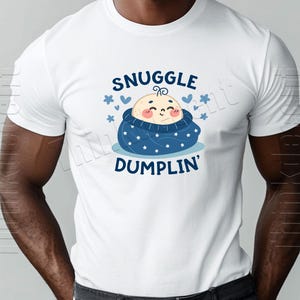 May include: White t-shirt featuring a cartoon baby wrapped in a blue blanket with white stars. The text "SNUGGLE DUMPLIN'" is above and below the baby illustration, with blue hearts and stars accents.