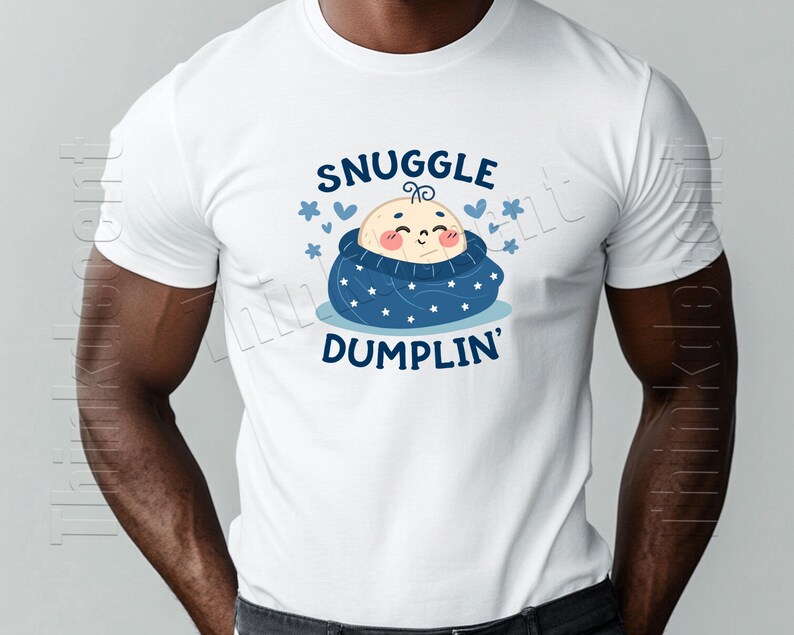 May include: White t-shirt featuring a cartoon baby wrapped in a blue blanket with white stars. The text "SNUGGLE DUMPLIN'" is above and below the baby illustration, with blue hearts and stars accents.