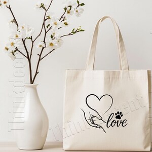 May include: Cream-colored tote bag with a black graphic of a hand holding a paw, a heart, and the word "love." A white vase with white flowers is to the left of the bag.