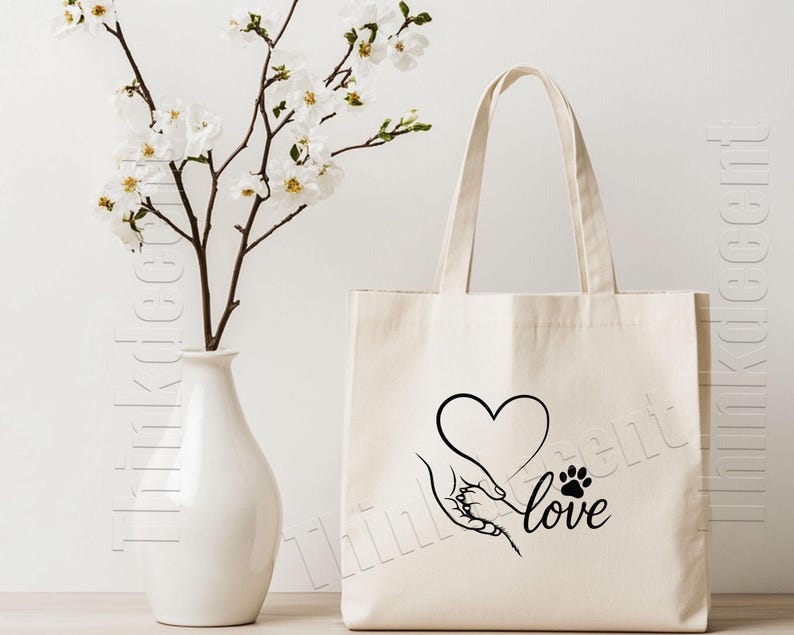 May include: Cream-colored tote bag with a black graphic of a hand holding a paw, a heart, and the word "love." A white vase with white flowers is to the left of the bag.