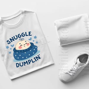 May include: White t-shirt with the text "SNUGGLE DUMPLIN'" and a cartoon baby design in blue and peach. A folded white cloth and a white sneaker are also in the image.
