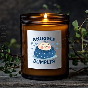 May include: A lit candle in a brown glass jar with a white label. The label features a cartoon baby wrapped in a blue blanket with the words "Snuggle Dumplin'" in blue. The candle sits on a wooden surface.