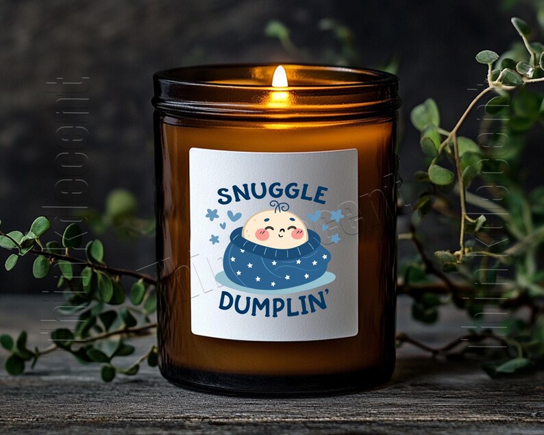 May include: A lit candle in a brown glass jar with a white label. The label features a cartoon baby wrapped in a blue blanket with the words "Snuggle Dumplin'" in blue. The candle sits on a wooden surface.