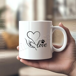 May include: White ceramic mug featuring a black graphic of a heart, a hand holding a paw, and the word "love." The mug has a curved handle and is held by a person. The design is simple and elegant, suitable for pet lovers.