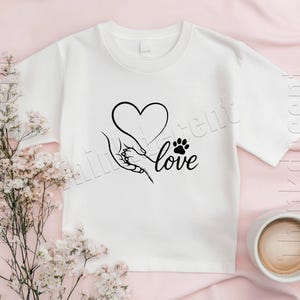May include: White t-shirt featuring a black graphic of a heart, a hand holding a paw, and the word "love." The design is centered on the shirt. A cup of coffee and flowers are in the background.