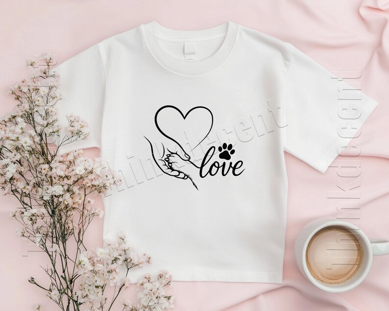 May include: White t-shirt featuring a black graphic of a heart, a hand holding a paw, and the word "love." The design is centered on the shirt. A cup of coffee and flowers are in the background.