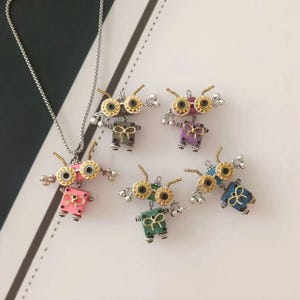 Op de afbeelding: Een verzameling kleurrijke robotvormige hangers met zilveren kettingen. Elke robot heeft een goudkleurige bril, armen en een strikje. De robots zijn roze, paars, groen, blauw en grijs. De hangers zijn waarschijnlijk sieraden.