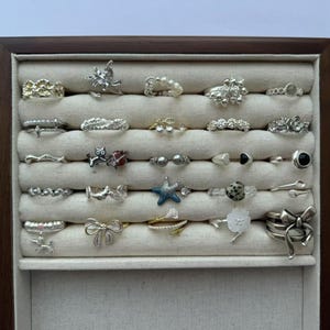 Puede incluir: Una exhibición de varios anillos en una caja de joyería de madera. Los anillos son plateados y presentan diversos diseños, incluyendo formas florales, de lazo y geométricas. La caja tiene un interior de tela clara.
