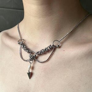May include: A silver chain necklace with a pearl and a silver spike pendant. The necklace has multiple chains and rings.