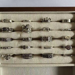 Puede incluir: Una caja de madera con forro de terciopelo, que muestra una colección de anillos de plata. Los anillos presentan varios diseños, incluyendo una corona, una cara sonriente y un corazón.