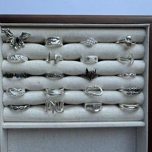 May include: A display case filled with various silver rings. The rings feature diverse designs, including geometric shapes, floral patterns, and abstract forms. The case has a wooden frame and a light-colored fabric interior, showcasing the rings against a neutral backdrop.