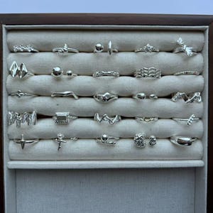 May include: A selection of silver rings displayed in a wooden-framed case. The rings showcase a variety of designs, from simple bands to more elaborate floral and geometric patterns. The interior of the case is a neutral colour.