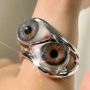 May include: A silver-toned metal ring featuring two realistic, three-dimensional eye designs. One eye has a blue iris, and the other has a brown iris. The eyes are set within a polished, curved metal band that wraps around the finger.