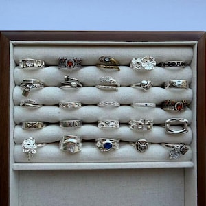 Puede incluir: Una vitrina llena de varios anillos de plata. Los anillos presentan diversos diseños, incluyendo detalles florales, geométricos y con piedras preciosas. La vitrina tiene un marco de madera y un interior de tela neutra, mostrando los anillos sobre un fondo suave.
