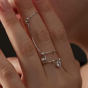 May include: A silver-toned double-band ring connected by a delicate box chain. The ring features a small polished heart charm and a round clear stone setting, with small spherical beads accenting the ends of the bands.
