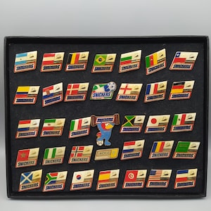 Könnte beinhalten: Eine Sammlung von Vintage-Snickers-Anstecknadeln, jede mit einem Flaggen-Design. Die Anstecknadeln sind in einem schwarzen Display-Koffer angeordnet. Die Flaggen repräsentieren verschiedene Länder, mit dem Snickers-Logo auf jeder Nadel.