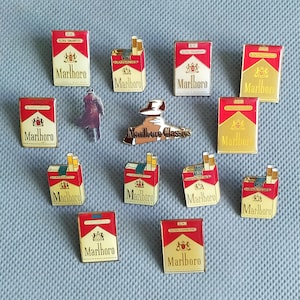 Könnte beinhalten: Eine Sammlung von dreizehn Metall-Emaille-Anstecknadeln mit verschiedenen Marlboro-Zigarettenpackungsdesigns in Rot, Weiß und Gold. Enthalten sind rechteckige Packungs-Pins, eine Figur im Trenchcoat und ein Pin mit dem Schriftzug Marlboro Classics.