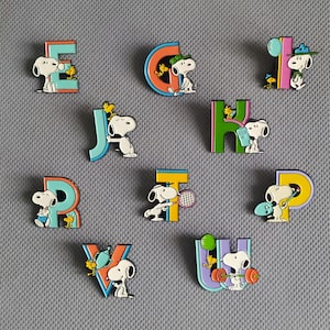 Peut inclure: Une collection de dix épinglettes en émail représentant le personnage Snoopy des Peanuts et son ami Woodstock interagissant avec diverses lettres colorées de l'alphabet. Chaque épinglette présente une scène unique, incluant des sports et loisirs.