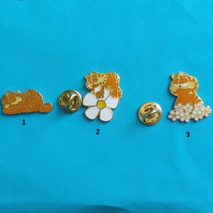 Peut inclure: Trois épingles en émail représentant le chat de dessin animé Garfield. Une épingle montre Garfield dormant, une autre Garfield sur une marguerite, et la troisième Garfield avec un groupe de points blancs. Les épingles sont orange, blanches et dorées.