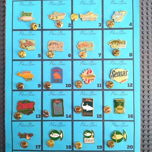 Puede incluir: Una colección de veinte pines de pesca Sensas sobre un fondo azul. Los pines presentan varios diseños de peces y el logotipo de Sensas. Cada pin tiene un respaldo dorado.