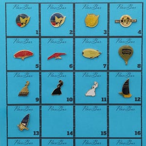 Puede incluir: Una colección de dieciséis pines de esmalte, cada uno con un diseño único. Los pines presentan varias formas, incluyendo un velero, un globo aerostático y figuras estilizadas. El fondo es una cuadrícula de cuadrados azules.