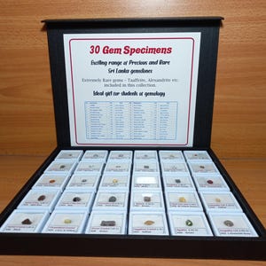 Może przedstawiać: Czarna gablota z 30 okazami kamieni szlachetnych. Gablota ma biały znak z czerwonym tekstem, kt&oacute;ry brzmi "30 Gem Specimens" i wymienia rzadkie kamienie szlachetne. Każdy kamień szlachetny jest prezentowany w małym, oznakowanym białym pudełku.
