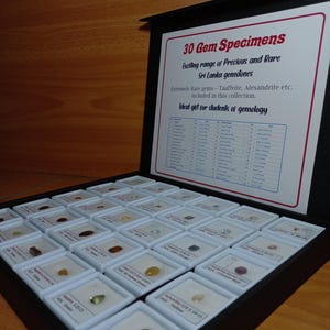 Może przedstawiać: Czarna gablota z 30 okazami kamieni szlachetnych, każdy w małym białym kwadracie. Gablota zawiera wydrukowaną kartę z tekstem "30 Gem Specimens" i "Exciting range of Precious and Rare Sri Lanka gemstones."