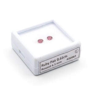 May include: A pair of small, round, pinkish-red rubies displayed inside a white square plastic storage box. A white label on the front of the box reads: Ruby Pair 0.62cts, Round 4.1 mm, #20983. The gemstones are positioned on a white foam insert.