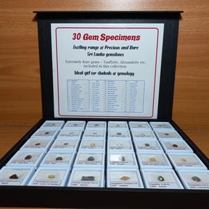 Może przedstawiać: Czarna gablota z 30 okazami kamieni szlachetnych. Gablota jest otwarta, ukazując małe, pojedynczo wyeksponowane kamienie szlachetne. Na g&oacute;rze gabloty znajduje się znak z napisem "30 Gem Specimens" i opisuje kolekcję.