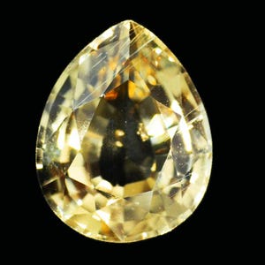 Puede incluir: Una piedra preciosa facetada en forma de pera con un tono amarillo. La piedra tiene un alto grado de claridad y se coloca sobre un fondo negro.