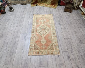 Puede incluir: Una alfombra rectangular de estilo vintage con un diseño geométrico central en tonos melocotón, beige y marrón. La alfombra tiene un aspecto desgastado, con una paleta de colores descoloridos y detalles intrincados en el borde. Está colocada sobre un suelo de madera clara.