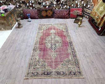 May include: A rectangular, vintage-style rug with a central medallion design. The rug features a faded, distressed look with a dominant pinkish-red hue, accented by cream and navy blue borders and details. The rug is displayed on a light wood floor.