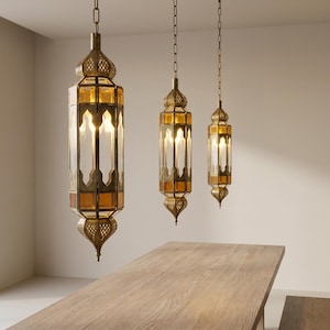 May include: Three ornate, gold-colored pendant lights with amber-colored glass panels hang from the ceiling. The lights have a traditional, lantern-like design with intricate metalwork and are suspended above a wooden table.