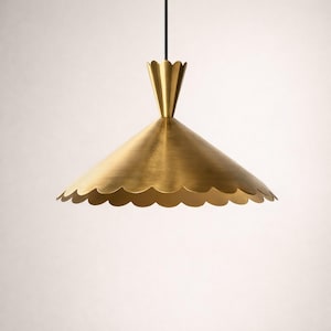 Könnte beinhalten: Eine messingfarbene Pendelleuchte mit gewelltem Rand. Die Leuchte hat einen konischen Schirm und eine dekorative Oberseite, die an einem schwarzen Kabel aufgehängt ist. Die Leuchte hat eine warme Goldfarbe und befindet sich vor einem weißen Hintergrund.