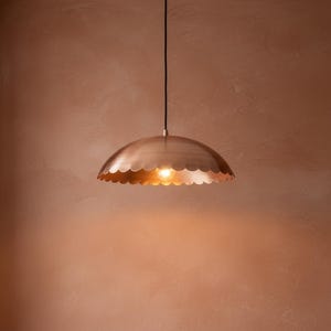 May include: A pendant light fixture with a scalloped edge, made of a warm-toned metal. The light hangs from a black cord and illuminates a soft glow. The fixture's design is reminiscent of a seashell.