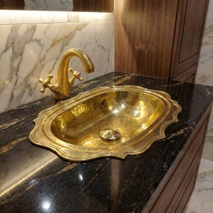 May include: A gold-coloured, hammered-metal sink with an ornate, scalloped edge. The sink is paired with a matching gold tap with cross-shaped handles. The sink is set on a black countertop with a dark wood cabinet.