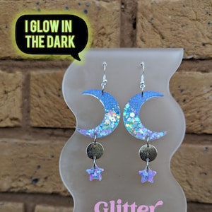 May include: A pair of crescent moon earrings with silver hooks and a star charm. The moon is made of clear resin with blue and iridescent glitter. The earrings glow in the dark. The earrings are displayed on a white stand with the text "Glitter Nonsense".