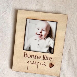 Può includere: Una cornice portafoto rettangolare in legno chiaro con un'apertura quadrata centrale contenente una fotografia in bianco e nero di un bambino sorridente. La cornice è incisa con il testo Bonne fête papa e un piccolo simbolo a forma di cuore.