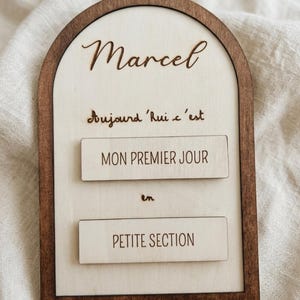 Peut inclure: Une plaque en bois en forme d'arche avec un cadre brun foncé et un centre en bois clair. Le texte indique : Marcel, Aujourd'hui c'est, MON PREMIER JOUR, en, PETITE SECTION. La plaque comporte deux inserts rectangulaires avec gravure noire.