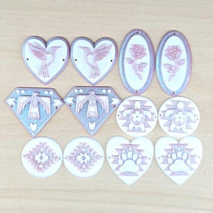 May include: A collection of twelve decorative craft components in various shapes including hearts, diamonds, ovals, and circles. Each piece features a white base with raised rose gold metallic designs such as hummingbirds, roses, eagles, turtles, geometric patterns, and bear paws.