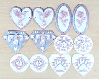 May include: A collection of twelve decorative craft components in various shapes including hearts, diamonds, ovals, and circles. Each piece features a white base with raised rose gold metallic designs such as hummingbirds, roses, eagles, turtles, geometric patterns, and bear paws.
