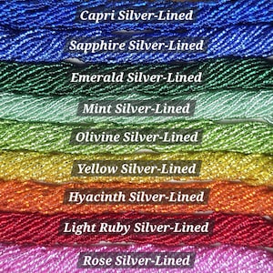 May include: A collection of eleven strands of silver-lined glass beads arranged horizontally. The colors include Straw Gold, Capri, Sapphire, Emerald, Mint, Olivine, Yellow, Hyacinth, Light Ruby, Rose, and Light Amethyst, each labeled with its name.