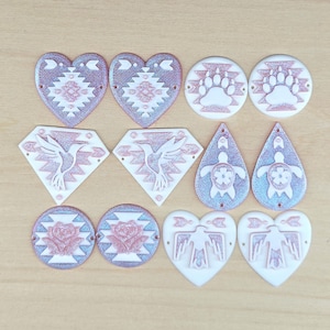 May include: A collection of ten flat, two-toned decorative charms in various shapes including hearts, circles, diamonds, and teardrops. Each piece features a raised relief design in white and shimmering rose gold, displaying motifs like hummingbirds, roses, turtles, bear paws, and birds.