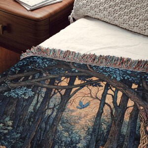 May include: A woven throw blanket featuring a detailed forest scene with dark blue and brown trees, a small blue bird in flight, and a light-colored sky. The blanket has a textured surface and a decorative fringe along the top edge with multi-colored threads.