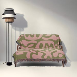 K&ouml;nnte beinhalten: Ein Zweisitzer-Sofa, das mit einer gewebten Decke mit olivgr&uuml;nem und altrosa abstraktem organischem Muster sowie einem mehrfarbigen Fransenrand bedeckt ist, neben einer hohen Stehlampe mit schwarz-wei&szlig;em Rattan-Lampenschirm.