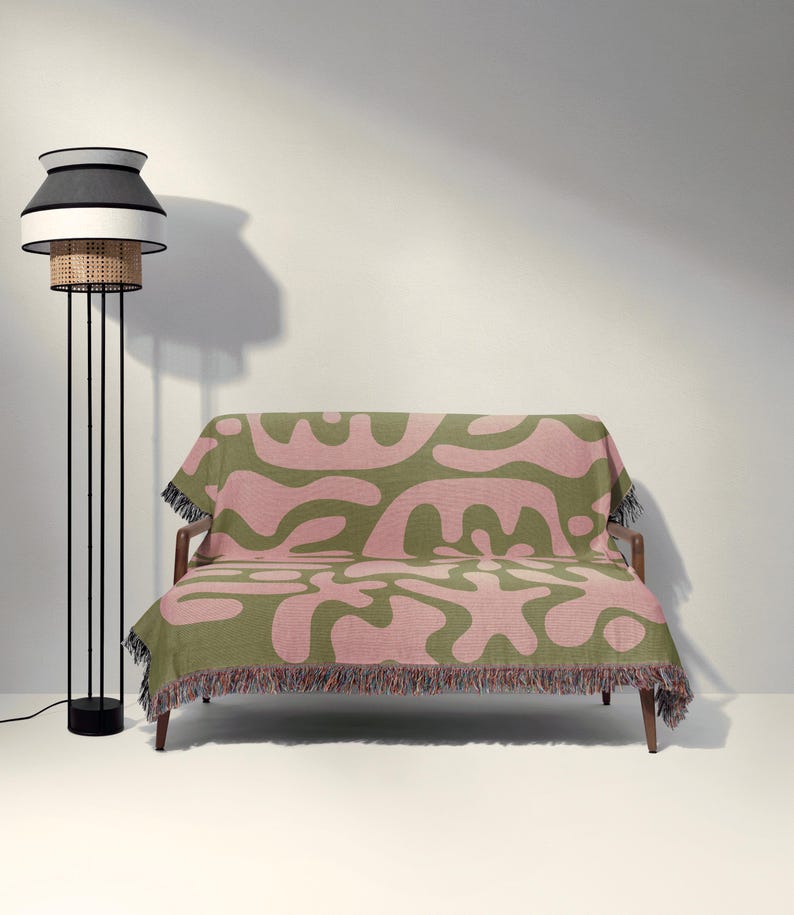 K&ouml;nnte beinhalten: Ein Zweisitzer-Sofa, das mit einer gewebten Decke mit olivgr&uuml;nem und altrosa abstraktem organischem Muster sowie einem mehrfarbigen Fransenrand bedeckt ist, neben einer hohen Stehlampe mit schwarz-wei&szlig;em Rattan-Lampenschirm.